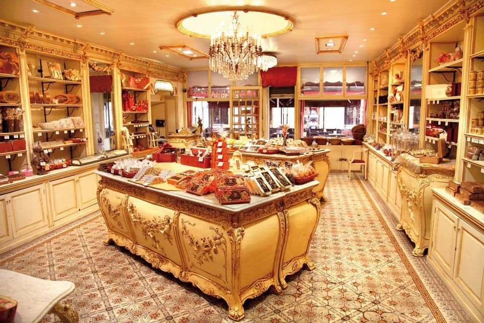 L'interno di una pasticceria sfarzosa in stile vintage, con banchi espositivi pieni di dolci e un lampadario di cristallo.