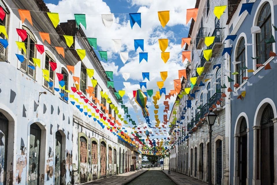 Banderas de festival coloridas y un farol de papel cuelgan sobre una calle estrecha de adoquines entre edificios antiguos bajo un cielo parcialmente nublado.