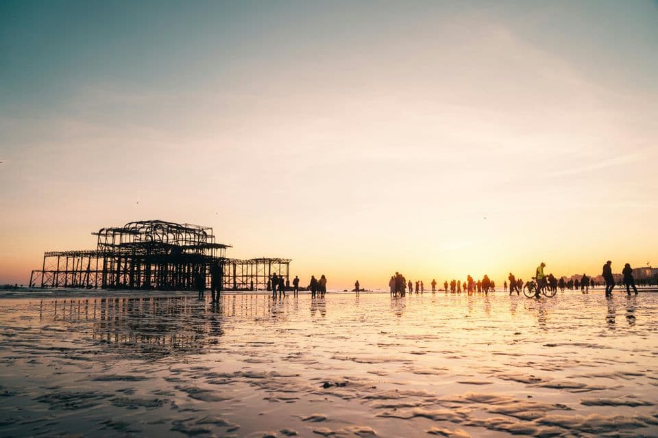 Sagome di persone su una spiaggia bagnata al tramonto, con la struttura scheletrica di un molo in rovina sullo sfondo.