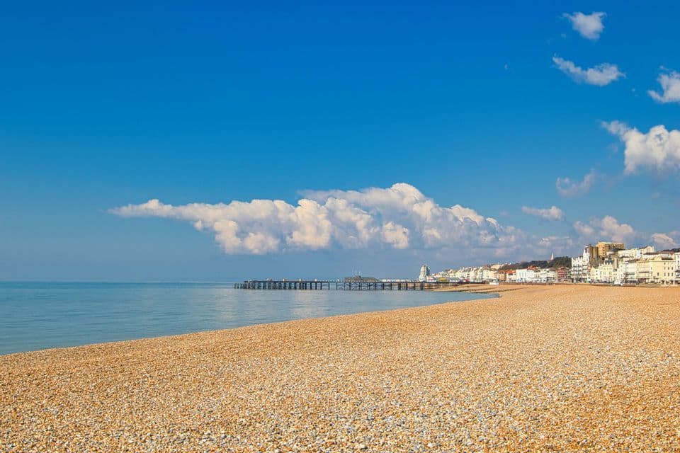 Una vasta spiaggia di ciottoli si incurva lungo un mare calmo verso un molo distante e una città costiera sotto un cielo azzurro brillante con nuvole bianche.