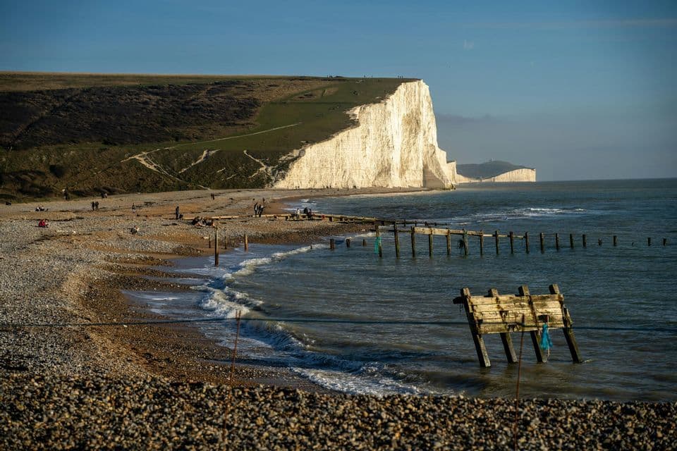 Ampia vista di una spiaggia di ciottoli con frangiflutti in legno che si estendono nel mare, sotto grandi scogliere bianche e un cielo soleggiato.