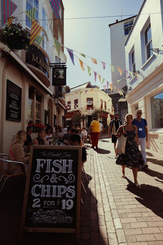 Una lavagna con l'offerta di fish and chips si trova su una strada di ciottoli, mentre le persone passeggiano e cenano in un pub all'aperto.