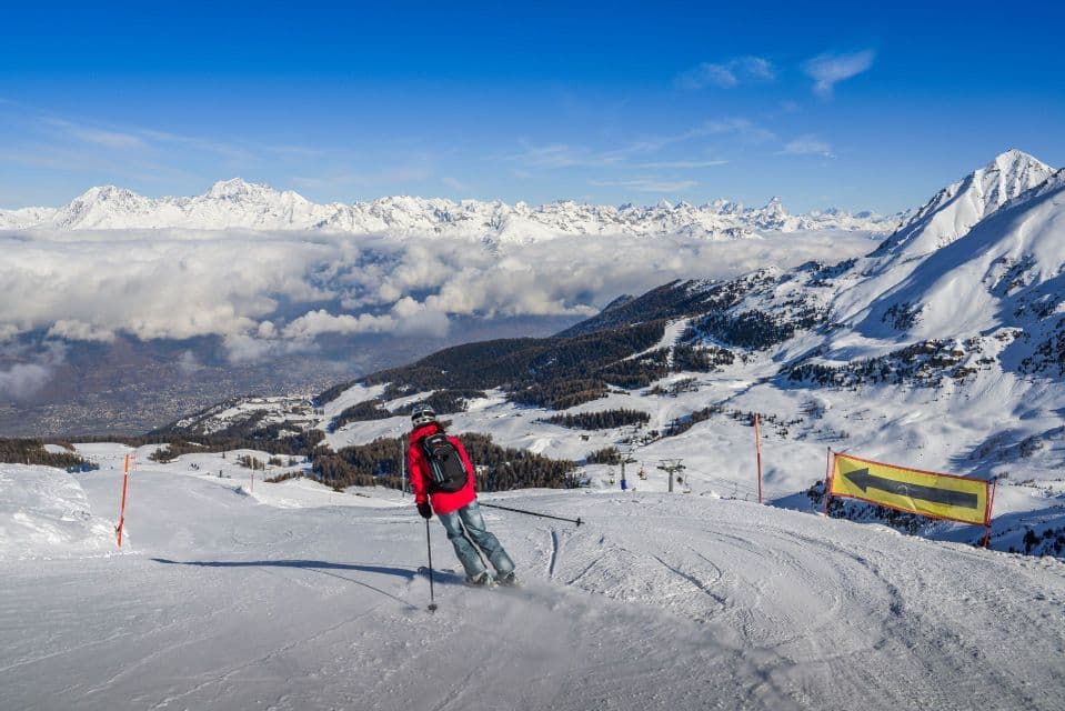 A skier in a red jacket descends a snowy mountain slope overlooking a sea of clouds and distant peaks under a clear blue sky.