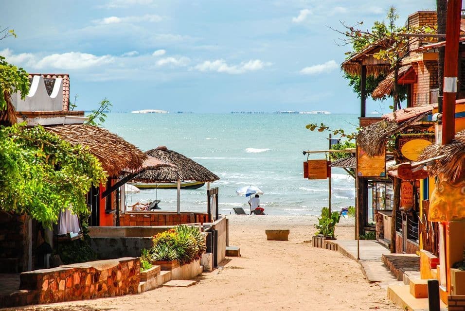 A sandy path between rustic buildings with thatched roofs leads down to a calm ocean beach under a partly cloudy sky.
