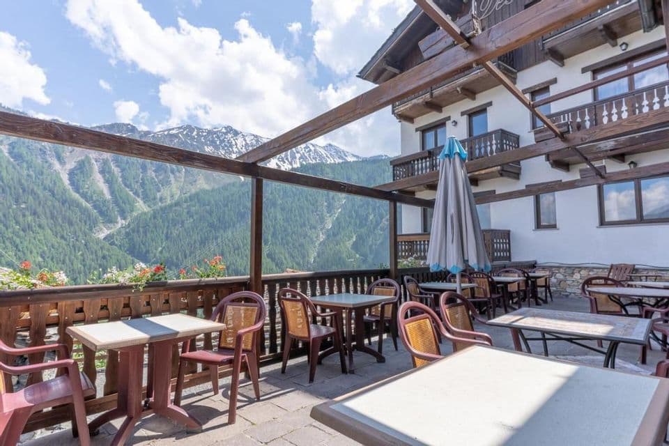 Una terrazza vuota di un hotel con tavoli e sedie sotto un pergolato, che si affaccia su una catena montuosa verde con cime innevate.