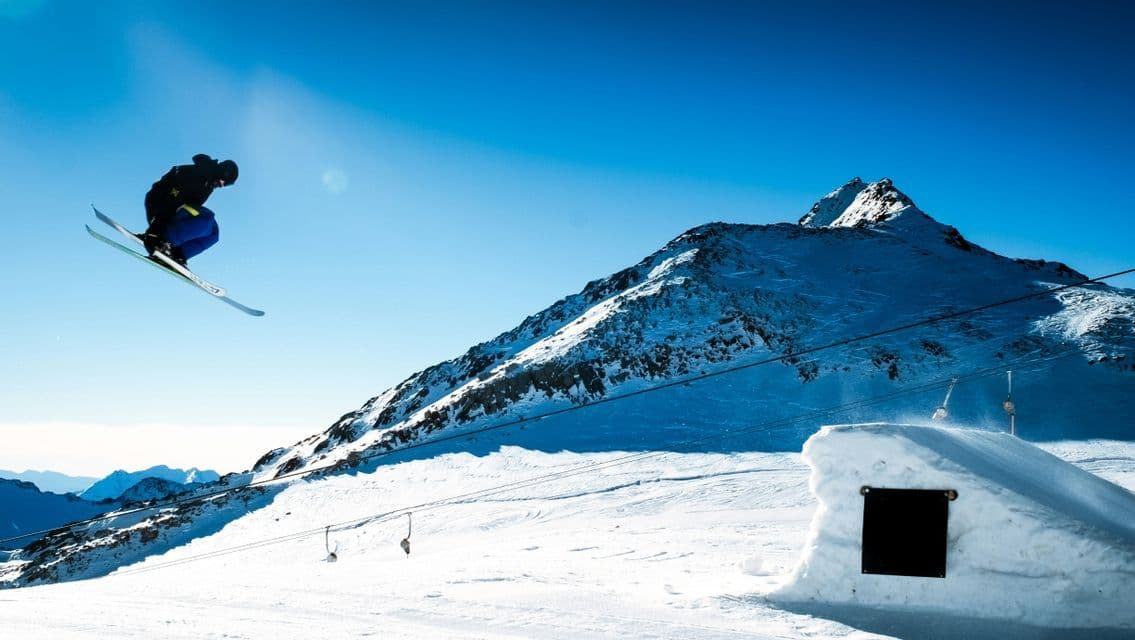 Uno sciatore è a mezz'aria mentre esegue un salto su una montagna innevata con un cielo azzurro limpido sullo sfondo.