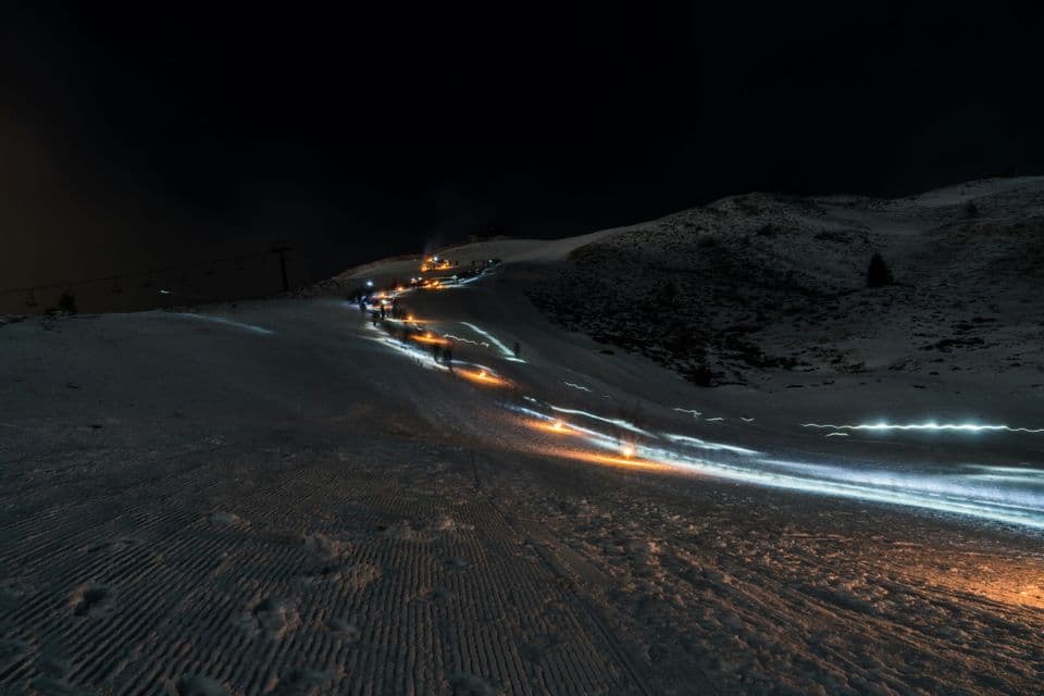 Uno scatto a lunga esposizione di un gruppo WeRoad in viaggio che sale un pendio innevato di notte, con le torce che creano scie di luce arancioni e bianche.