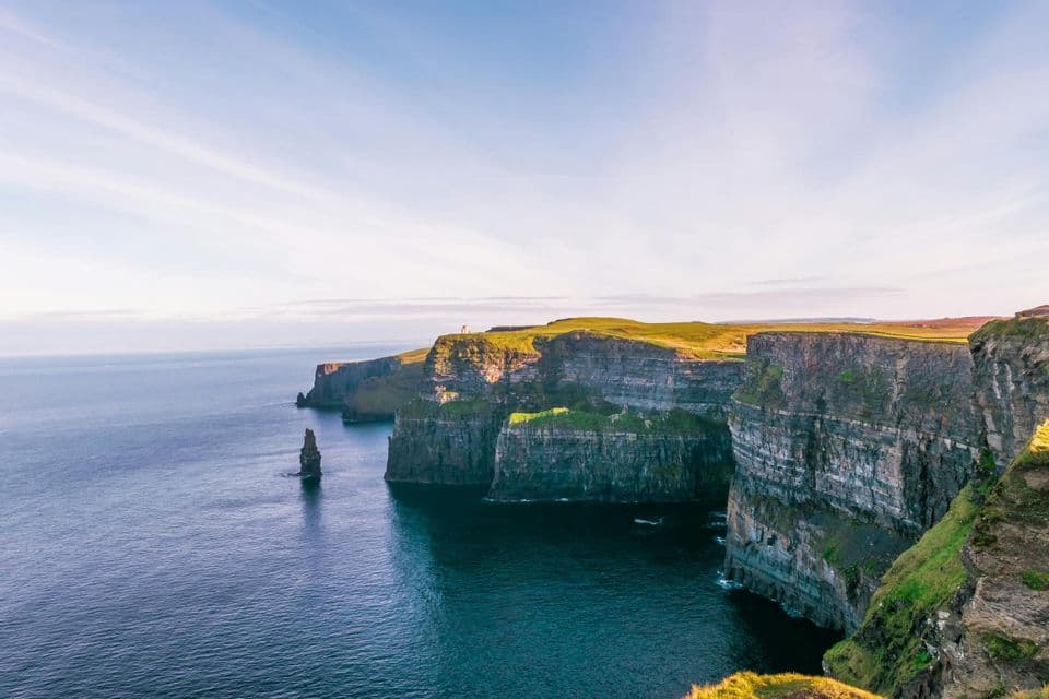 D'abruptes falaises aux sommets verdoyants surplombent un océan bleu et calme, avec une haute aiguille rocheuse sous un ciel dégagé.