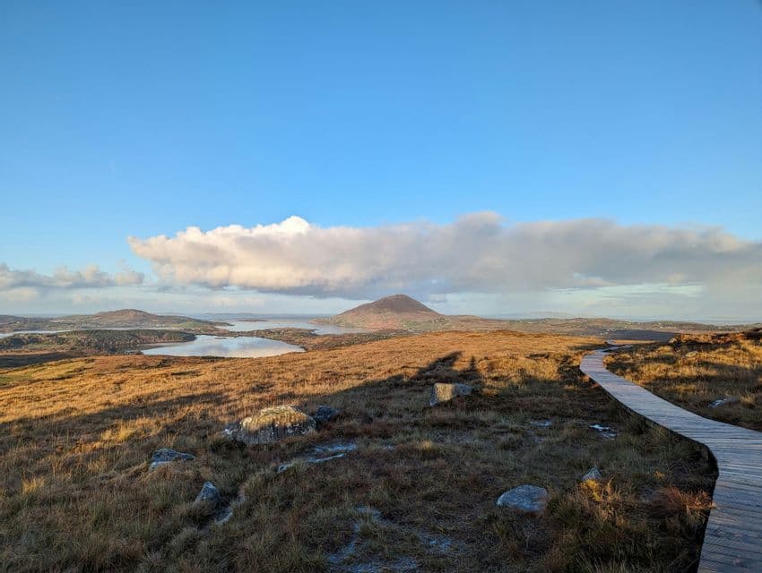 Une promenade en bois serpente à travers un vaste paysage herbeux avec des montagnes et des lacs au loin sous un ciel bleu.