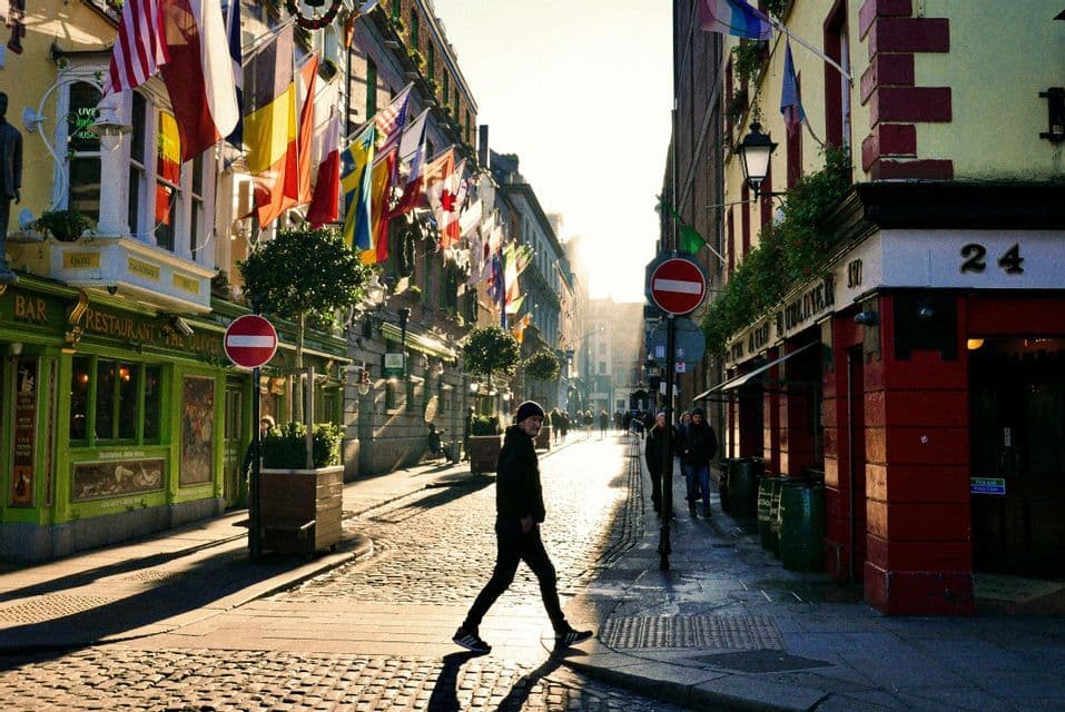 Une personne traverse une rue pavée ensoleillée bordée de bâtiments ornés de drapeaux internationaux.