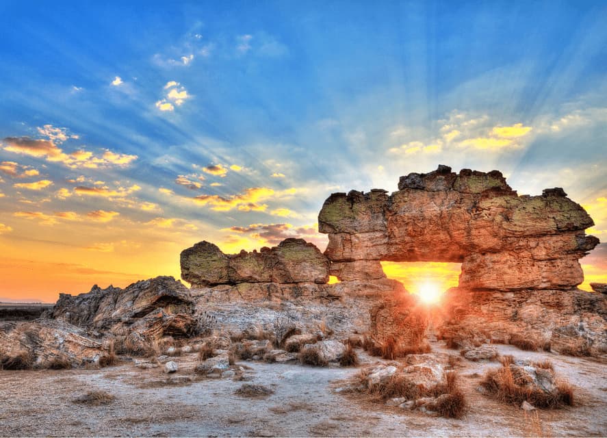 The sun shines through a large natural rock arch formation at sunset, casting rays across a blue and yellow sky.