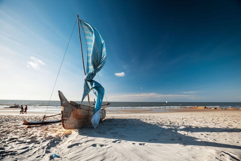 Ein Holzboot mit einem aufgerollten blau-weißen Segel liegt an einem weißen Sandstrand und wirft einen langen Schatten unter einem klaren blauen Himmel.