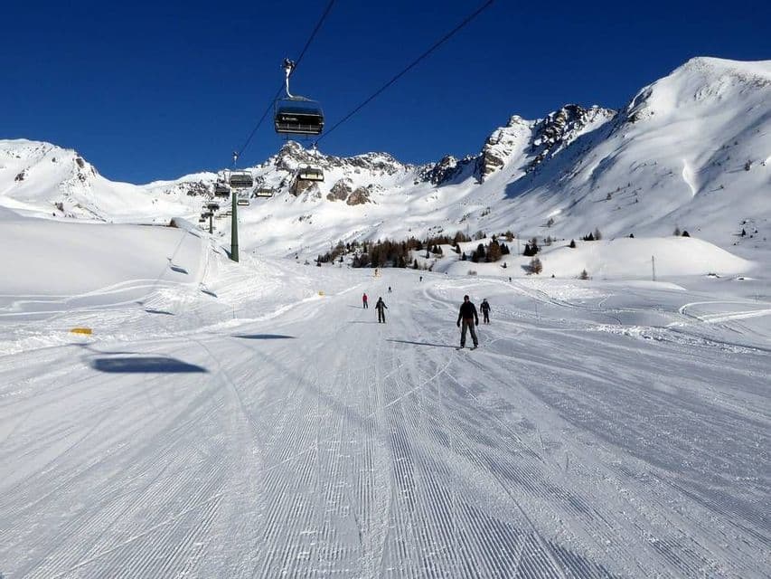 Diverse persone sciano su un'ampia pista battuta sotto una seggiovia in una giornata di sole, con le montagne innevate sullo sfondo.