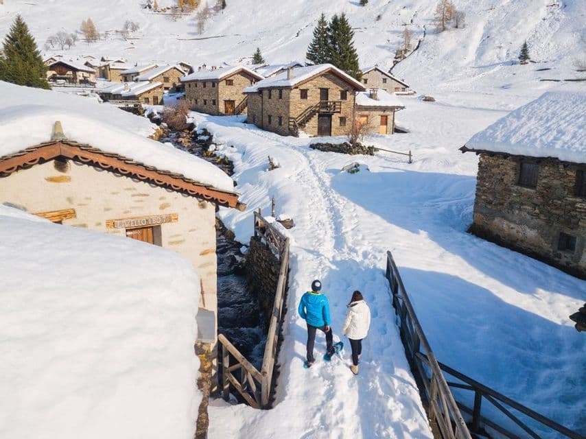 Vista dall'alto di due persone con le ciaspole che camminano su un sentiero attraverso un villaggio di montagna innevato con case in pietra.