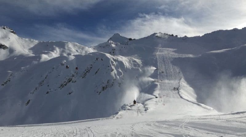 Cannoni da neve in funzione su una pista da sci in un vasto paesaggio montano innevato con seggiovie sotto un cielo blu.