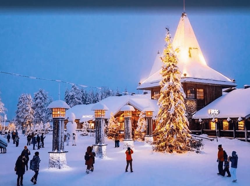 Al crepuscolo, persone passeggiano in una piazza di paese innevata, illuminata da un grande albero di Natale, lanterne ed edifici coperti di neve.