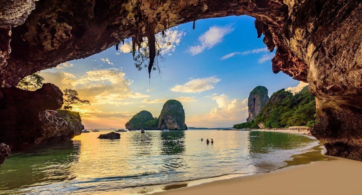 Una vista desde el interior de una cueva marina con vistas a una playa de arena, kársticos de piedra caliza y personas en el agua durante un atardecer dorado.