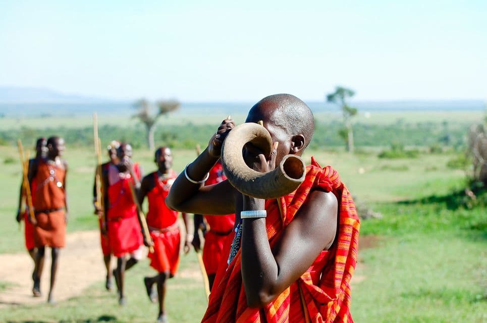A Maasai man in red traditional clothing blows a horn while other tribesmen walk behind him in a grassy savanna.
