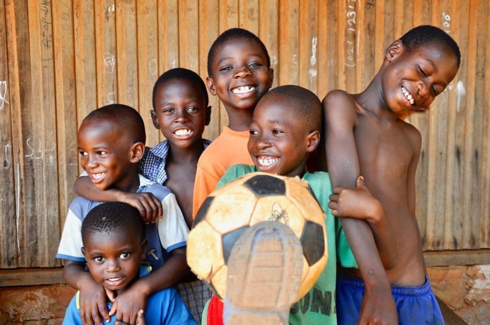Un grupo de seis niños pequeños sonriendo juntos frente a una pared de madera, uno de ellos sosteniendo un balón de fútbol gastado.