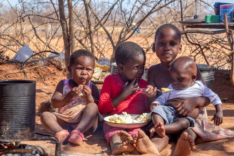 Cuatro niños pequeños se sientan juntos en el suelo al aire libre, compartiendo una comida de un solo plato.