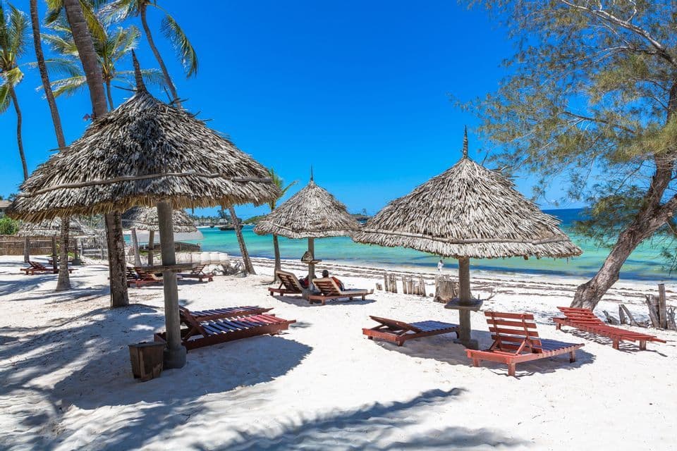 Strohdächer und hölzerne Liegestühle an einem weißen Sandstrand mit Palmen und türkisfarbenem Wasser unter einem klaren blauen Himmel.