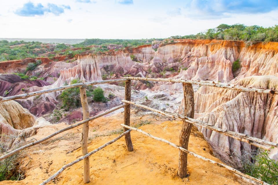 Una recinzione di legno su un belvedere sabbioso offre una vista su un grande canyon con formazioni di arenaria colorate ed erose sotto un cielo blu.