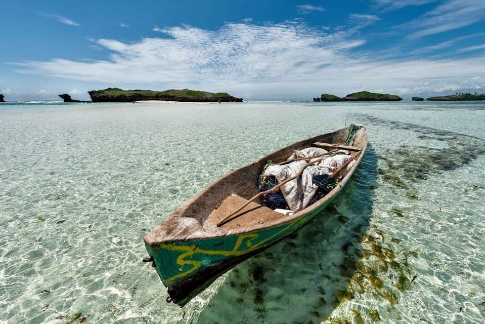 Una canoa de madera verde con aparejos de pesca flota en aguas poco profundas y cristalinas, con pequeñas islas en el horizonte bajo un cielo azul.