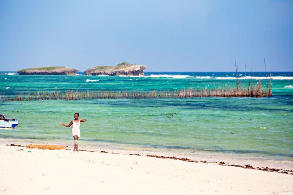 Une jeune fille en robe claire se tient sur une plage de sable blanc, au bord d'une eau turquoise, avec des îlots rocheux en arrière-plan.