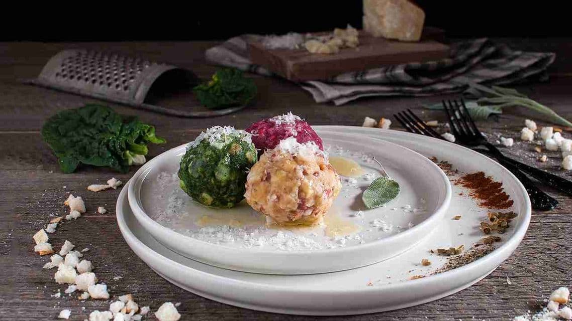 Un piatto di tre gnocchi colorati—verdi, rossi e beige—guarniti con formaggio grattugiato e una foglia di salvia su un tavolo di legno.
