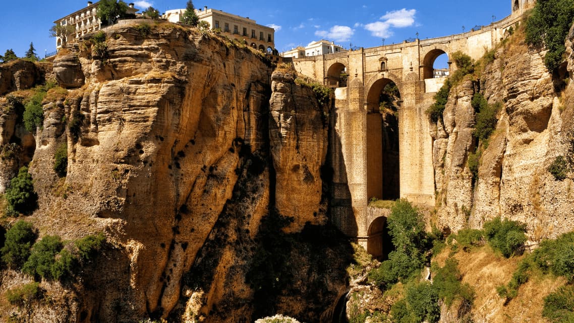 A large stone bridge with multiple arches spans a deep, rocky gorge with buildings perched on the clifftop under a blue sky.