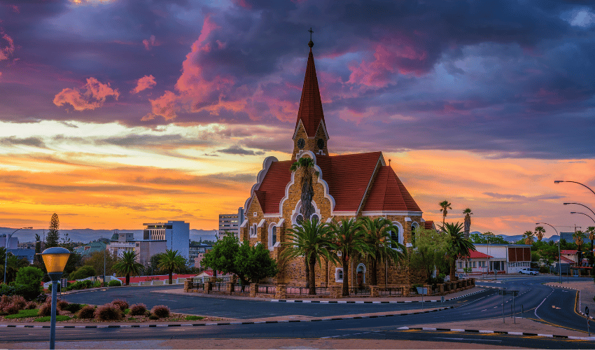 Una chiesa in pietra con tetto rosso e campanile su uno spartitraffico cittadino sotto un cielo drammatico al tramonto viola e arancione.