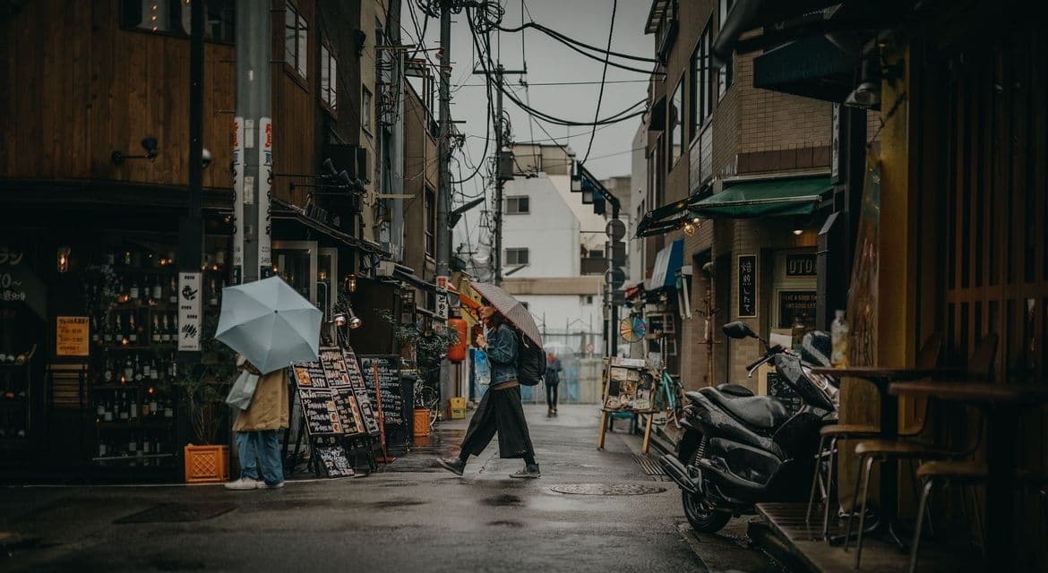 Persone con ombrelli camminano attraverso un vicolo stretto e bagnato di città, fiancheggiato da negozi e fili aerei aggrovigliati.