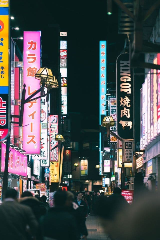 Una folla di persone cammina lungo una strada stretta di notte, illuminata da alte insegne al neon luminose con caratteri giapponesi.