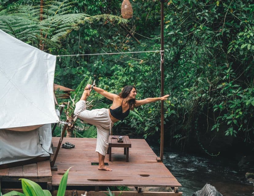 Una donna mantiene una posizione yoga su una pedana di legno accanto a una tenda bianca, con una giungla e un fiume sullo sfondo.