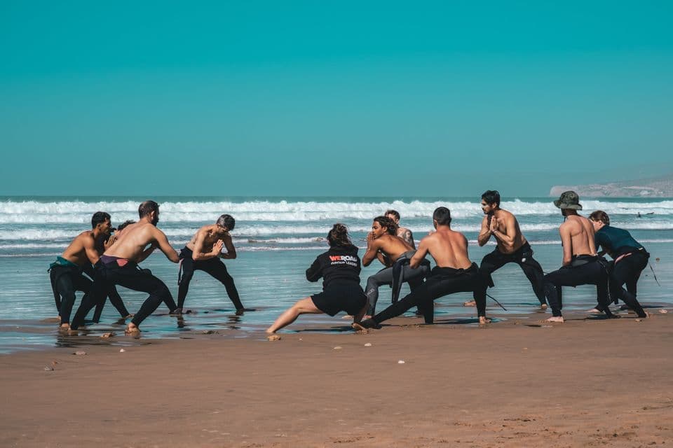 Un gruppo WeRoad in viaggio che fa stretching di riscaldamento sulla spiaggia, con le onde dell'oceano sullo sfondo.