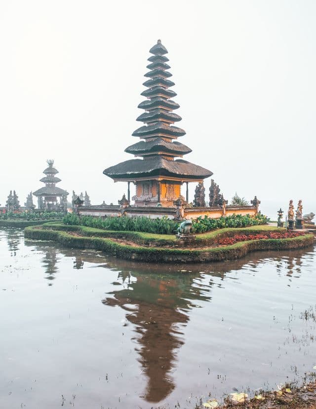 Un tempio tradizionale a più livelli sorge su una piccola isola, il cui riflesso si specchia sull'acqua calma sotto un cielo nebbioso.
