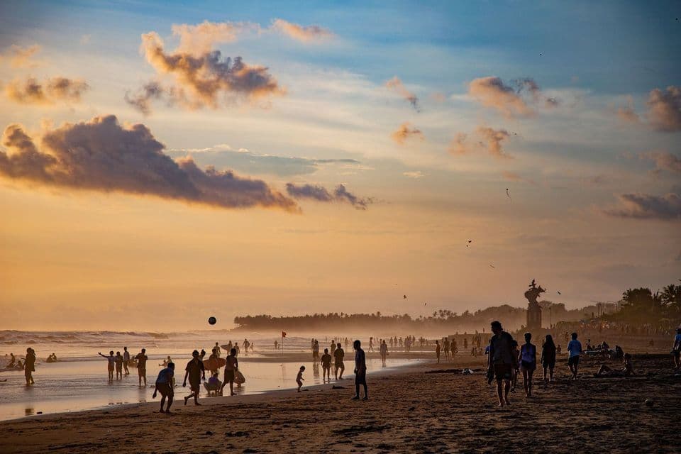 Una folla di persone in silhouette su una spiaggia sabbiosa al tramonto, che camminano e giocano in riva al mare.