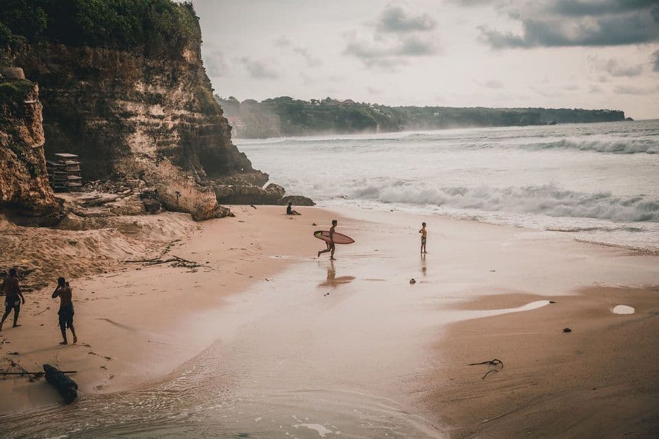 Un surfista attraversa una spiaggia sabbiosa portando la tavola verso l'oceano, con una grande scogliera rocciosa sulla costa.
