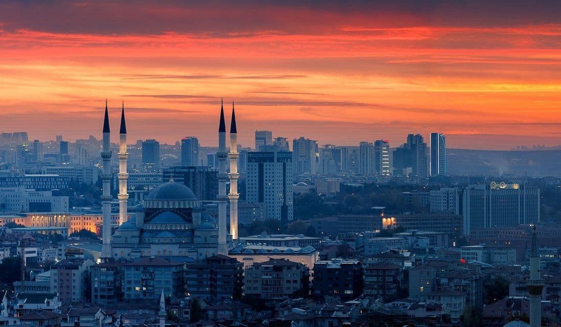 Una grande moschea con minareti illuminati e una cupola si erge in un paesaggio urbano denso sotto un cielo al tramonto dai colori vivaci arancioni e rossi.