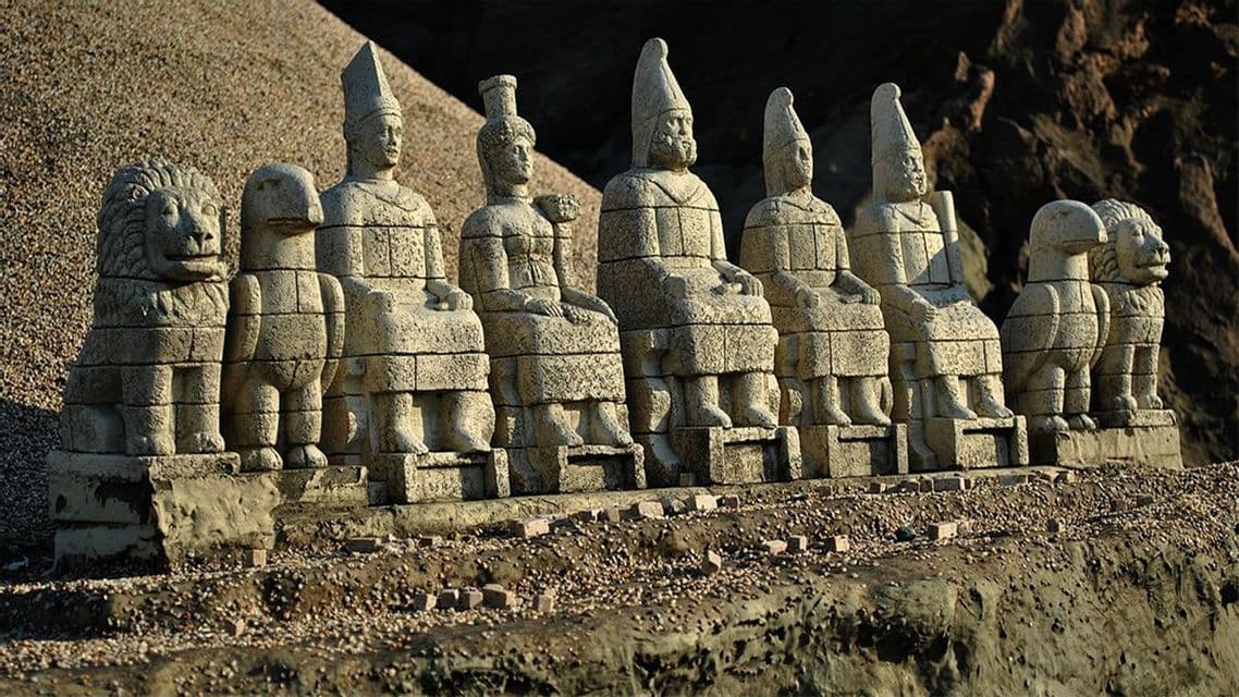 Una fila di grandi statue di pietra sedute raffiguranti figure antiche, affiancate da sculture di leoni e aquile, su una sporgenza ghiaiosa.
