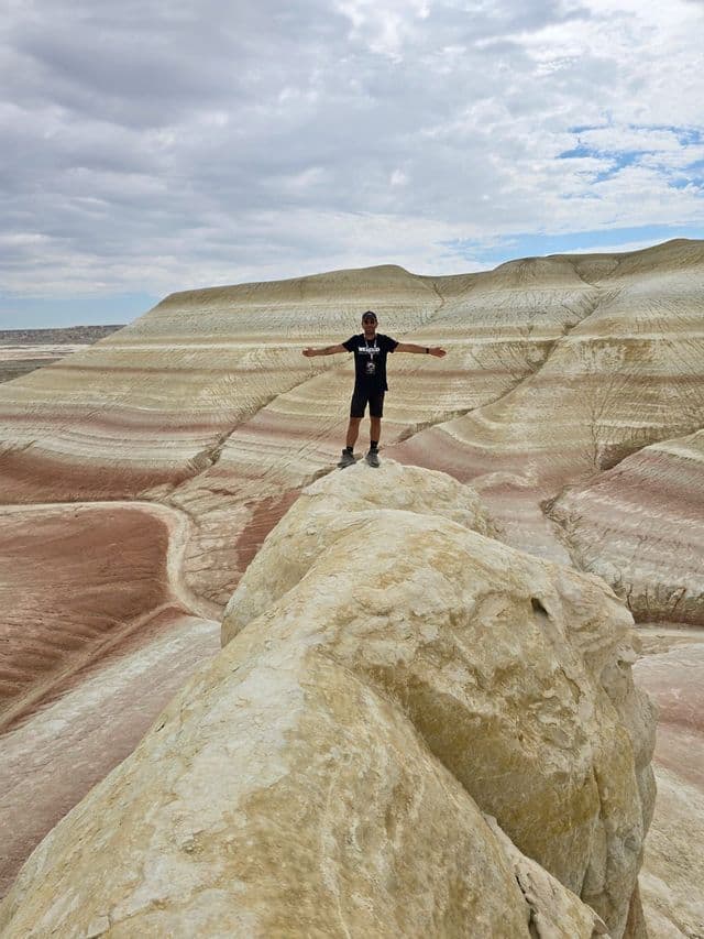 Un uomo si erge a braccia aperte su una formazione rocciosa, ammirando un canyon di colline colorate e striate sotto un cielo nuvoloso.