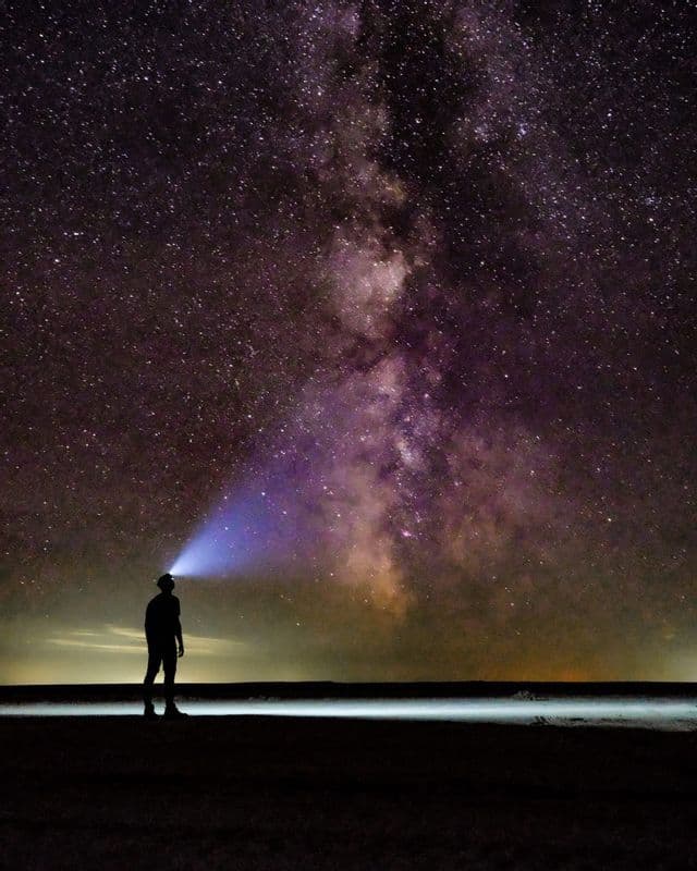Une personne en silhouette portant une lampe frontale lève les yeux vers la Voie lactée sous un vaste ciel nocturne rempli d'étoiles.