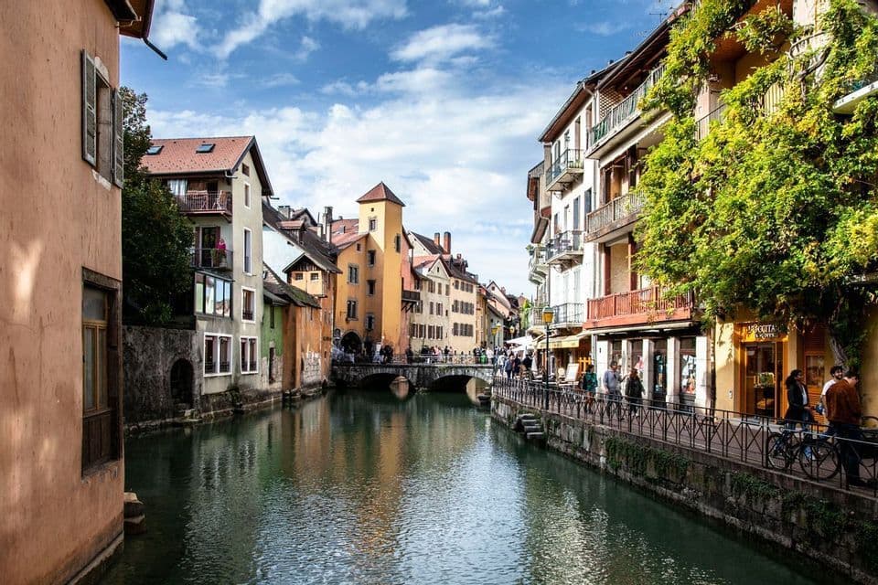 A canal flows between colorful, historic buildings in a European town as people walk along the waterfront and over a stone bridge.