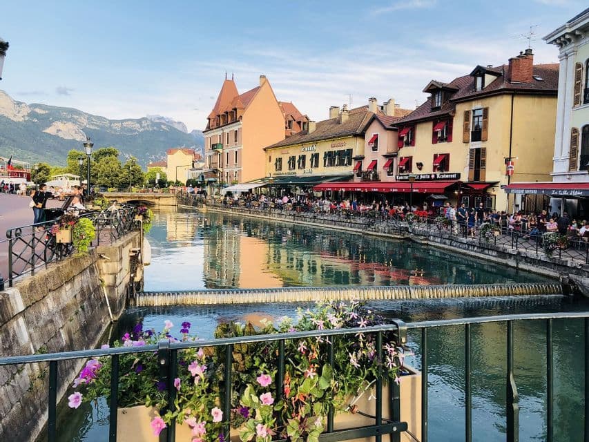 A canal flows through a town with colorful buildings and restaurants with outdoor seating, with mountains visible in the background.