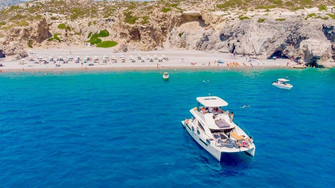 Veduta aerea di un viaggio di gruppo WeRoad su un catamarano in una baia turchese, con una spiaggia sabbiosa affollata e scogliere in lontananza.