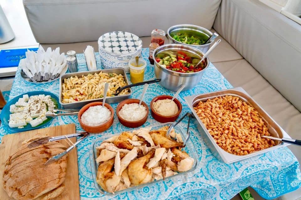 Un pasto a buffet con vari piatti tra cui pasta, pollo, insalata e pane, disposti su un tavolo con una tovaglia blu.