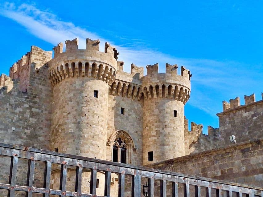 Una fortezza medievale in pietra con due grandi torri merlate contro un cielo azzurro brillante, vista oltre un cancello scuro.
