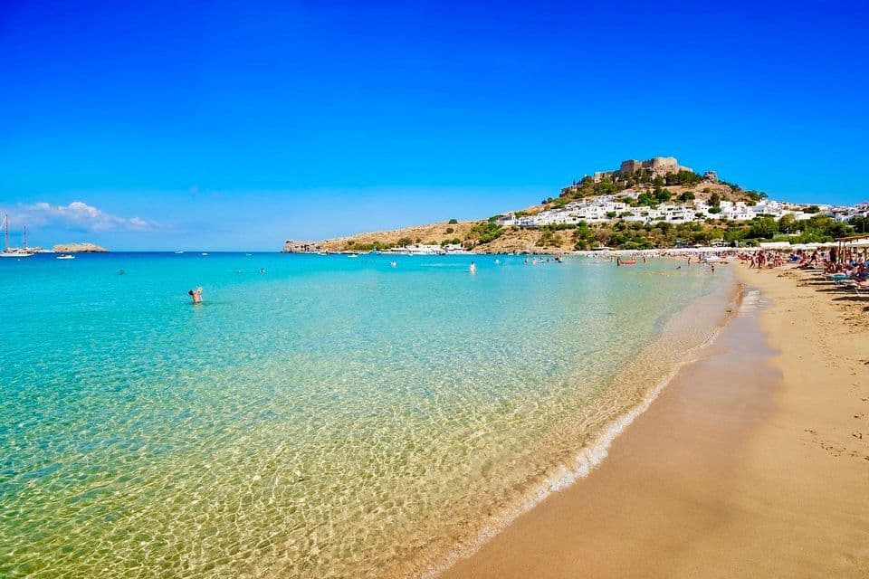 Una spiaggia sabbiosa con acqua cristallina color turchese, gente che nuota e un borgo collinare con una fortezza sullo sfondo.