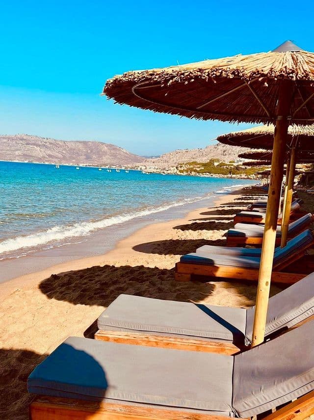 Una fila di lettini vuoti sotto ombrelloni di paglia su una spiaggia sabbiosa affacciata su un mare turchese e colline distanti.