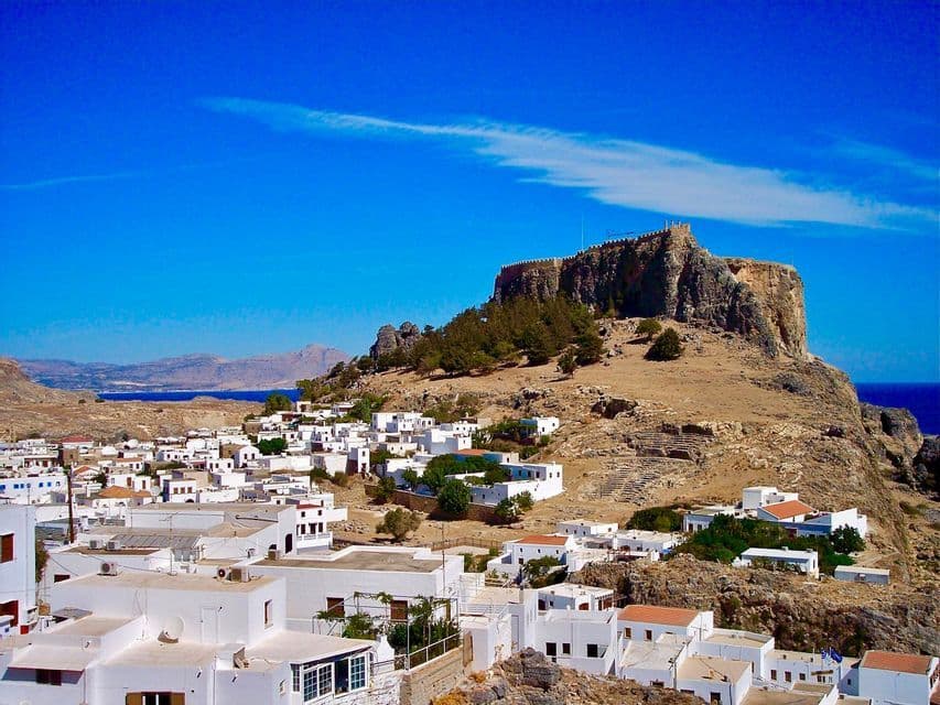 Un'antica acropoli sorge su una collina rocciosa che domina un villaggio di case bianche e il mare sotto un cielo azzurro e limpido.